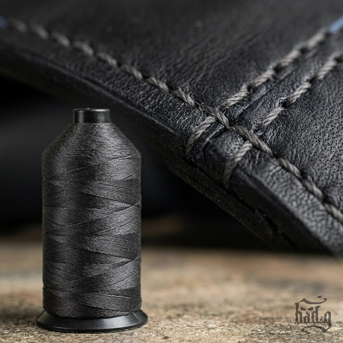 Spool of gray thread next to a piece of black leather with stitching.