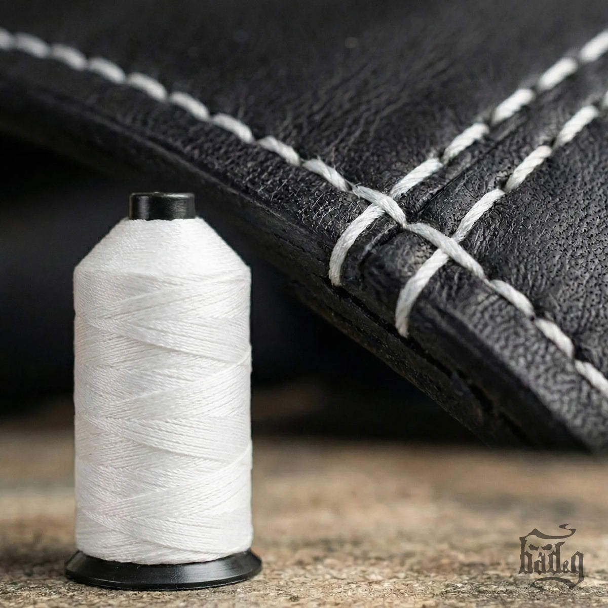 Spool of white thread next to a black leather product with white stitching on a wooden surface.